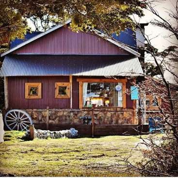 The image depicts a charming red cabin with a corrugated metal roof, surrounded by a stone wall and lush greenery, exuding a cozy and rustic atmosphere.