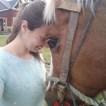 The image depicts a woman standing beside a brown horse, with the horse\'s head positioned in front of her. The woman is wearing a light blue sweater.