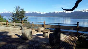 A peaceful wooden deck overlooks a serene lake, with rustic log chairs and table. Snow-capped mountains and clear blue skies create a tranquil, natural setting.