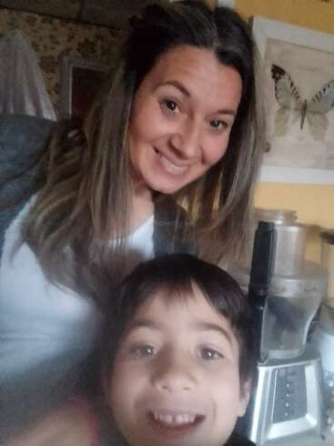 A smiling woman and a cheerful child pose for a selfie in a cozy kitchen, with a butterfly picture and a food processor visible in the background.