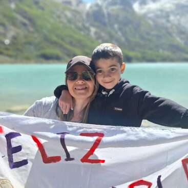 A woman and a young boy smile together by a beautiful turquoise lake, holding a colorful sign. Mountains and greenery form a picturesque natural background.