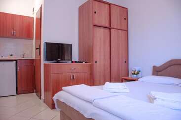 Das Bild zeigt ein Zimmer mit einem Bett, einem Tisch und einem Stuhl, mit einem großen Fenster und einer Tür, mit einer weißen Wand und einem grauen Boden, die eine einfache und saubere Atmosphäre schaffen.