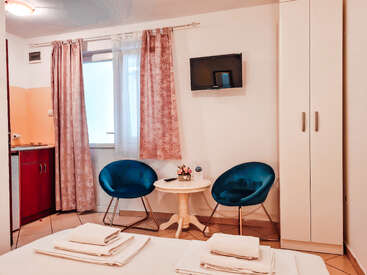 Das Bild zeigt ein kompaktes Hotelzimmer mit einem Bett, zwei Stühlen, einem Tisch und einem Fernseher, mit weißen Wänden und einem Fenster mit rosa Vorhängen.