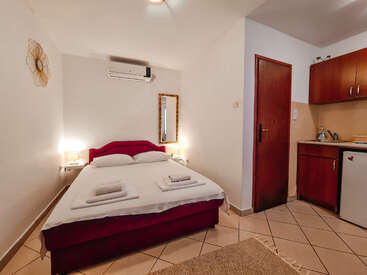 Das Bild zeigt ein kompaktes Schlafzimmer mit Bett, Spiegel und Küchenzeile, das mit seinen weißen Wänden, dem gefliesten Boden und dem roten Bettgestell eine gemütliche Atmosphäre ausstrahlt.
