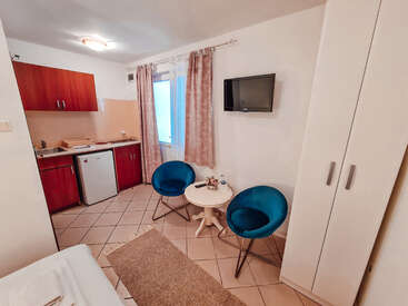 Das Bild zeigt eine kompakte Studiowohnung mit einer Küchenzeile, zwei blauen Stühlen, einem kleinen Tisch und einem an der Wand montierten Fernseher, mit weißen Wänden und gefliestem Boden.
