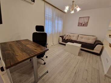 Das Bild zeigt einen kleinen, gut beleuchteten Raum mit einem Schreibtisch, einem Stuhl, einer Couch und einem Couchtisch. Weiße Wände, Holzböden und ein Kronleuchter schaffen eine gemütliche Atmosphäre.