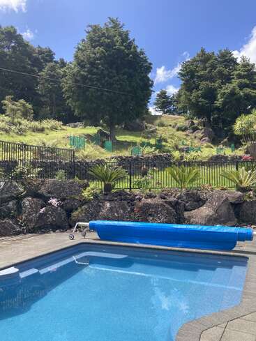 Im Vordergrund liegt ein klarer, blauer Swimmingpool, der von Felsen, Pflanzen und einem schwarzen Zaun umgeben ist, dahinter liegen grüne Hügel und hohe Bäume.