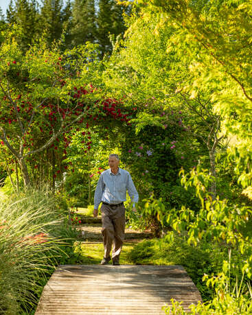 Ein älterer Mann spaziert über eine Holzbrücke in einem üppigen Garten. Er ist umgeben von sattem Grün und blühenden Blumen, was eine friedliche und ruhige Atmosphäre schafft.