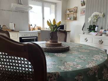 Une cuisine chaleureuse à la décoration florale. Une table ronde, une nappe verte à motifs floraux, un centre de table composé de tulipes jaunes, des chaises en bois, une fenêtre lumineuse et des meubles blancs créent un espace accueillant.