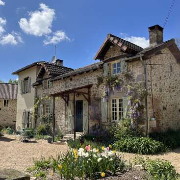 La imagen representa una encantadora casa de piedra con tejado de tejas, rodeada de exuberante vegetación y flores vibrantes, bajo un sereno cielo azul.