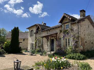 L'image représente une charmante maison en pierre avec un toit en tuiles, entourée d'une végétation luxuriante et de fleurs éclatantes, sur fond de ciel bleu serein.
