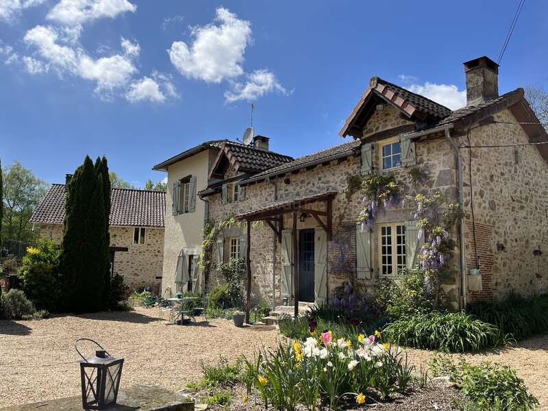 L'image représente une charmante maison en pierre avec un toit en tuiles, entourée d'une végétation luxuriante et de fleurs éclatantes, sur fond de ciel bleu serein.