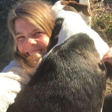 The image shows a woman with blonde hair and a white fleece jacket, smiling and hugging a large dog with a brown and white coat.
