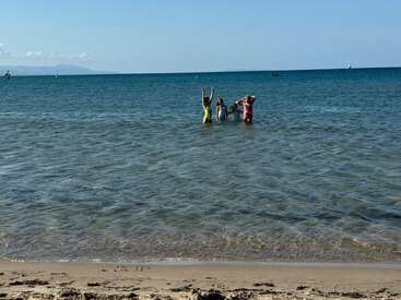Three children stand in the shallow sea, playing and raising their arms joyfully, while the clear blue water and sandy beach create a peaceful summer scene.