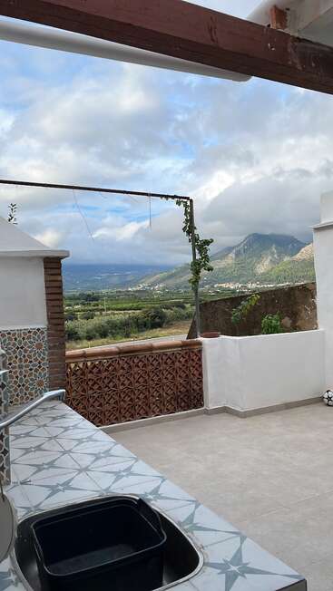 A tiled outdoor kitchen with a sink overlooks a beautiful green valley and mountains under a partly cloudy sky. Decorative tiles and a soccer ball add charm.