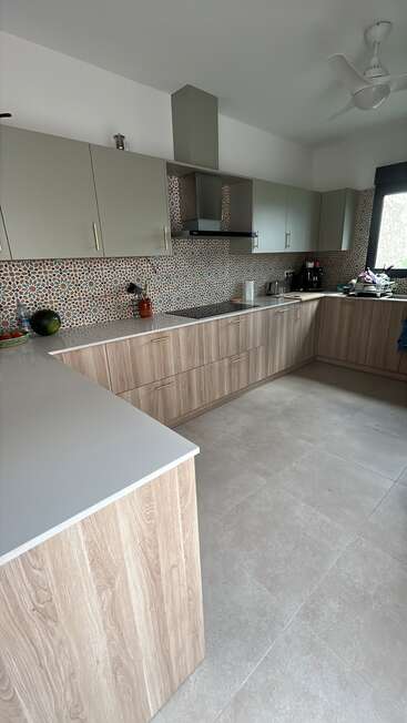 This kitchen features light wood cabinets, grey countertops, patterned tile backsplash, modern appliances, a ceiling fan, and ample natural light through a large window. Cozy and stylish.