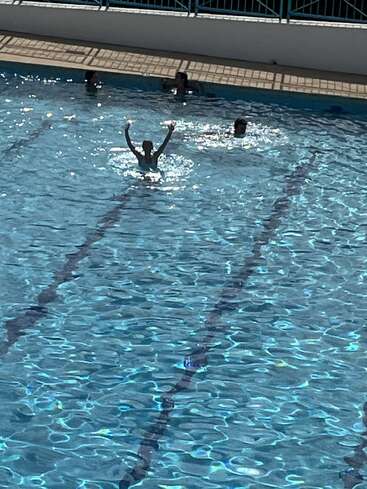 In a swimming pool, several people are enjoying the water. One person stands out, raising both arms triumphantly while the sunlight sparkles on the surface.