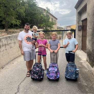 A man and four children, three with rolling backpacks, stand on a paved lane by a gate, smiling. It's a bright, casual outdoor scene.