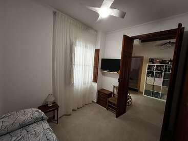 A small, cozy bedroom with a single bed, side table, lamp, sheer curtains, ceiling fan, wall-mounted TV, wooden chair, and open doorway to another room.