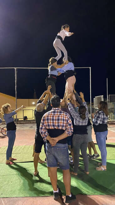 A group of people are forming a human tower outdoors at night, supporting each other as a young girl carefully climbs to the top, balancing confidently.