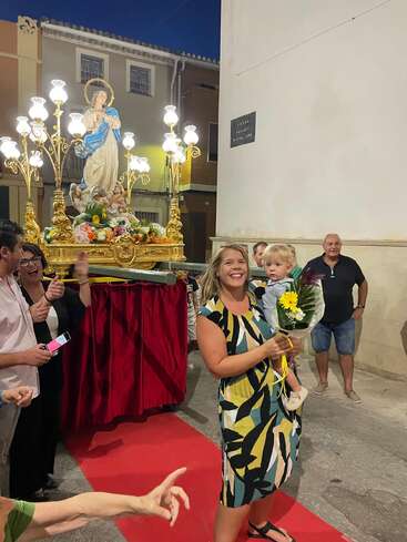 A smiling woman holds a small child and bouquet, surrounded by people at a festive nighttime event with a religious statue adorned with lights and flowers.