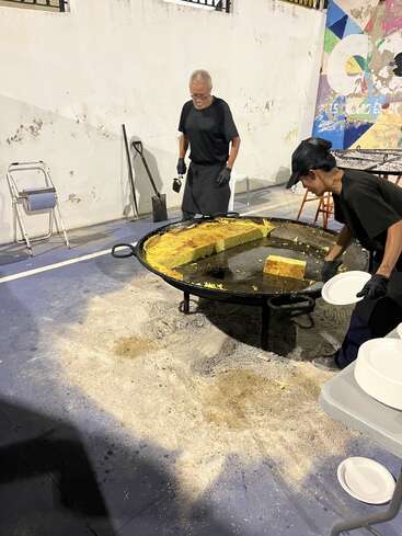 Two people are preparing food outdoors using a giant pan containing a large yellow rectangular dish. Plates are stacked nearby, and the ground is covered in sawdust.