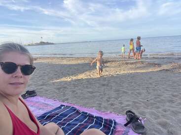A woman in sunglasses sits on a beach towel, while children play by the shoreline under a blue sky. Sandals and another towel are nearby.