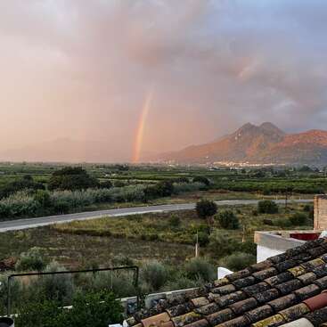 Un arc-en-ciel éclatant apparaît au-dessus de champs verdoyants et de routes sinueuses, avec des montagnes en arrière-plan et un ciel dramatique et nuageux au-dessus, vu d'un toit rustique.