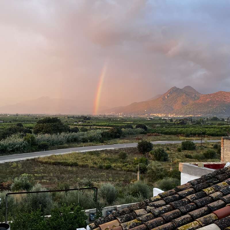 A vibrant rainbow appears over green fields and winding roads, with mountains in the background and dramatic, cloudy skies above, viewed from a rustic rooftop.