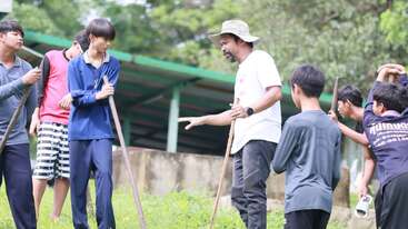A group of young people and an adult man are outdoors, holding sticks. The adult appears to be instructing or teaching them, possibly about gardening or teamwork.