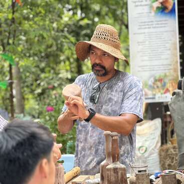 The image depicts a man wearing a straw hat, standing behind a table with various wooden objects, possibly engaged in a demonstration or workshop.