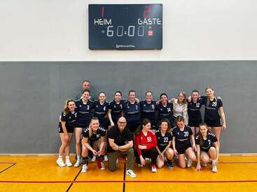 Un equipo femenino de balonmano posa bajo un marcador que indica 2-1. Parecen felices, juntas en una pista cubierta, celebrando tras su partido.