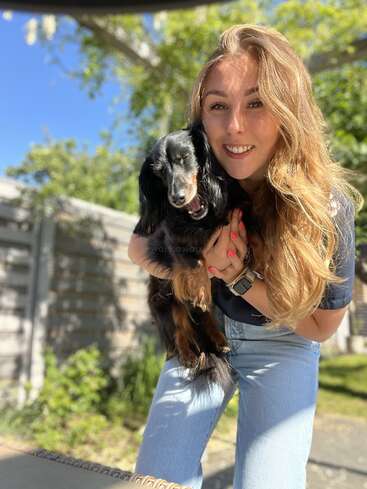 Une jeune femme aux longs cheveux blonds sourit en tenant un petit chien noir à l'extérieur par une journée ensoleillée, avec de la verdure et une clôture en bois derrière eux.