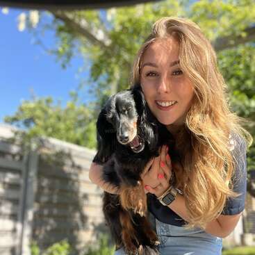 Une jeune femme aux longs cheveux blonds sourit en tenant un petit chien noir à l'extérieur par une journée ensoleillée, avec de la verdure et une clôture en bois derrière eux.