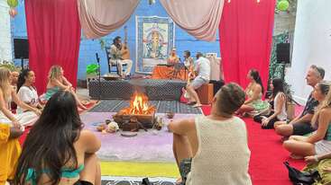 Um grupo de pessoas senta-se em círculo ao redor de uma fogueira em um ambiente fechado, participando de uma sessão espiritual ou musical com artistas, decorações vibrantes e uma atmosfera pacífica.
