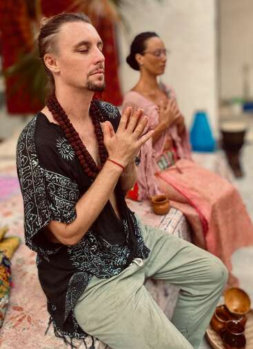 Duas pessoas estão sentadas com os olhos fechados e as mãos em posição de oração, parecendo meditar. Elas usam roupas e acessórios descontraídos, cercadas por xícaras e decoração colorida.
