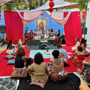 A group of people sits on colorful cushions around a stage where musicians perform. Vibrant decorations, red drapes, and lanterns create a warm, festive atmosphere.