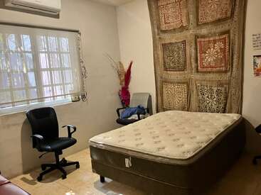 Um quarto aconchegante com um colchão, cadeiras, tapeçaria de retalhos na parede, janela com persianas, ar-condicionado e flores secas decorativas no canto.