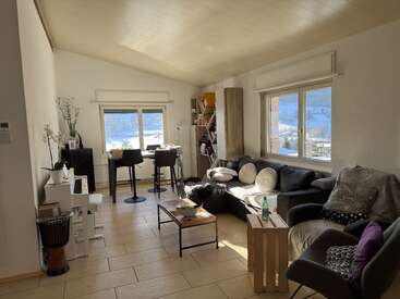 This cozy, sunlit living room features a black sofa, modern furniture, a piano, bookshelf, and large windows offering a scenic snowy mountain view outside. Warm tones throughout.