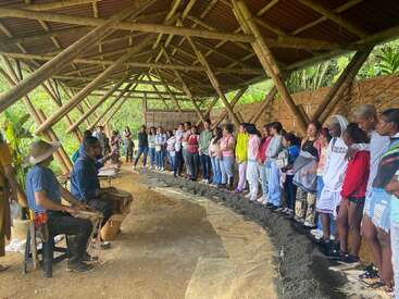 A group of people stand in a semi-circle under a rustic bamboo shelter, listening attentively to two seated presenters with drums. The setting appears natural.
