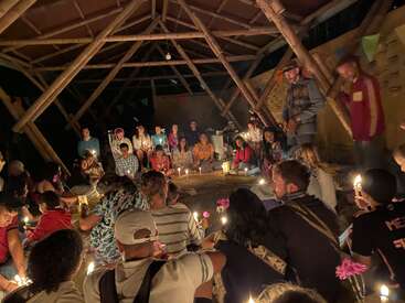 Un groupe de personnes est assis en cercle à l'intérieur d'une structure en bambou, chaleureusement éclairée par des bougies, créant une atmosphère intime, paisible et communautaire lors d'une réunion nocturne.
