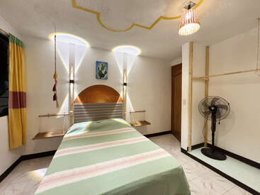 Cette chambre est équipée d'un lit simple recouvert d'un couvre-lit à rayures vertes, d'un éclairage lumineux, d'une décoration minimaliste, de rideaux jaunes, d'étagères murales, d'un ventilateur sur pied et d'un plafond au design original.