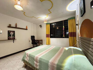 Une chambre confortable avec un couvre-lit rayé, des rideaux jaunes et verts, un plafond au design original, des décorations murales, un bureau, une chaise, des suspensions et des étagères décoratives ornées d'objets colorés.