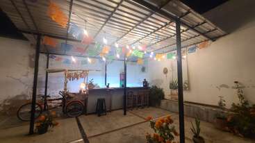Une terrasse extérieure festive est agrémentée de banderoles colorées en papel picado, de guirlandes lumineuses, d'un petit bar, de tabourets, de soucis, d'un tricycle décoré et de plantes en pot.