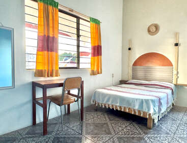 Une chambre lumineuse et sobre, avec un lit orné d'une couverture décorative, un bureau et une chaise en bois près de la fenêtre, des rideaux colorés, un sol carrelé et un chapeau accroché au mur.