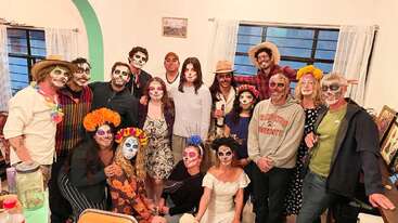 A imagem mostra um grupo de pessoas reunidas, adornadas com pinturas faciais vibrantes e trajes coloridos, posando para uma foto em uma sala com paredes brancas e uma grande janela.