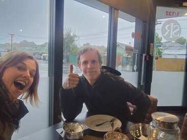Zwei Leute sitzen an einem Restauranttisch und lächeln begeistert in die Kamera. Einer streckt den Daumen hoch, vor ihnen sind Essen und Besteck zu sehen.
