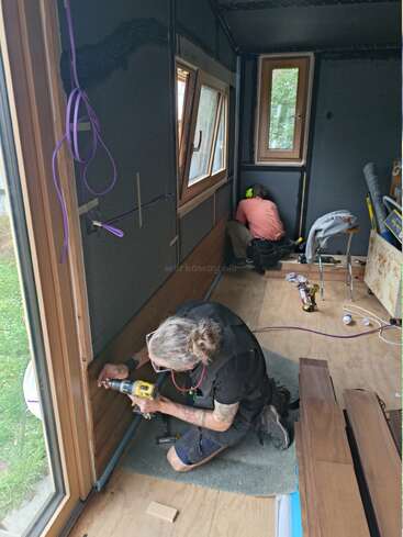 In einem kleinen Raum, der gerade umgebaut wird, arbeiten zwei Leute. Der eine bohrt in der Nähe eines Fensters Löcher in Holzplatten, während der andere sich in der Ecke um die Verkabelung kümmert.