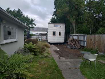 Am Ende eines Gartenwegs steht ein kleines weißes Tiny House. Farne, Bäume, ein Holzzaun und Gartenstühle umrahmen diese friedliche Hinterhofszene.