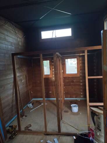 Ein kleiner Raum aus Holz, der gerade im Bau ist, mit freiliegenden Wandkonstruktionen, Werkzeugen, Kabeln und verstreutem Material. Drei Fenster sorgen für Tageslicht. Der Boden und die Wände sind noch unfertig.
